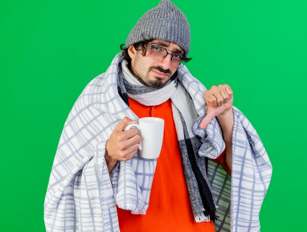 안경 겨울 모자와 스카프를 착용하는 불쾌한 젊은 백인 아픈 남자가 녹색 벽에 고립 된 아래로 엄지 손가락을 보여주는 격자 무늬 지주 컵에 싸여
