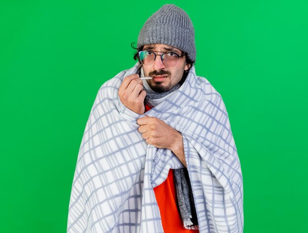 Unpleased young caucasian ill man wearing glasses winter hat and scarf wrapped in plaid putting thermometer in mouth  grabbing plaid isolated on green wall with copy space