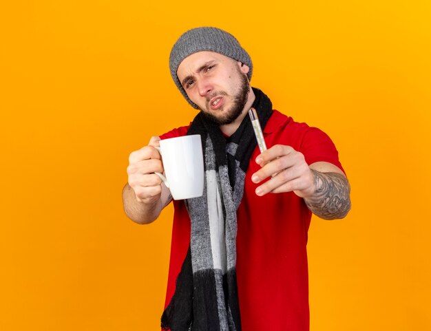 Unpleased young caucasian ill man wearing winter hat and scarf holding cup and thermometer isolated on orange wall with copy space