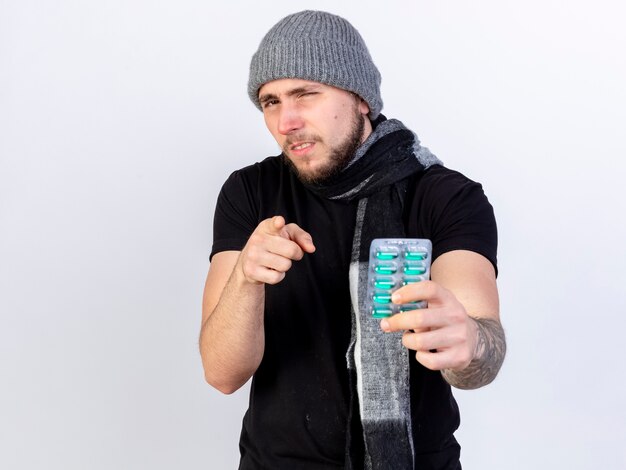 Unpleased young caucasian ill man wearing winter hat and scarf holds pack of medical capsules and points isolated on white wall with copy space