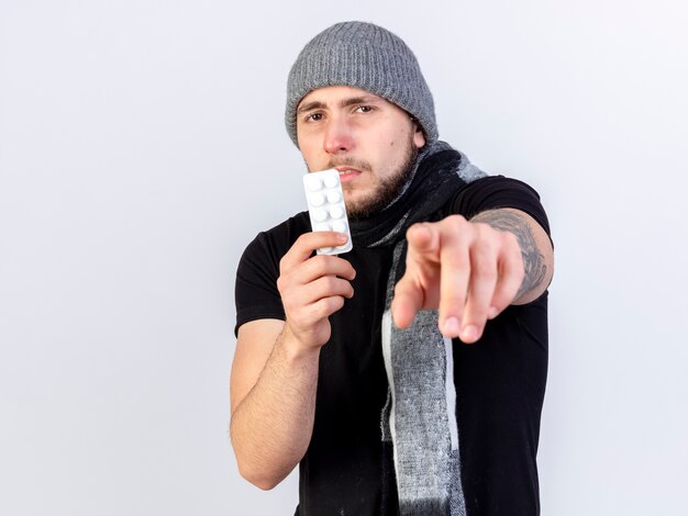Unpleased young caucasian ill man wearing winter hat and scarf holds pack of medical tablets pointing isolated on white wall with copy space