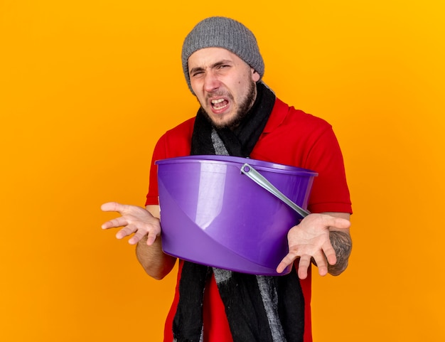 Unpleased young caucasian ill man wearing winter hat and scarf holds plastic bucket isolated on orange wall with copy space