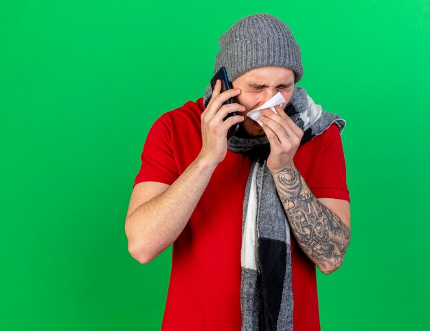 冬の帽子とスカーフを身に着けている不機嫌な若い白人の病気の人は、緑の電話で話しているティッシュで鼻を拭きます
