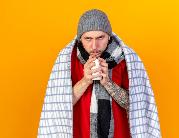 Unpleased young caucasian ill man wearing winter hat and scarf wrapped in plaid holds cup isolated on orange wall with copy space