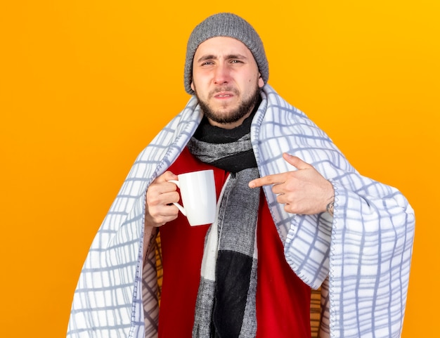 格子縞のホールドとコピースペースでオレンジ色の壁に隔離されたカップを指す冬の帽子とスカーフを身に着けている不機嫌な若い白人の病気の男