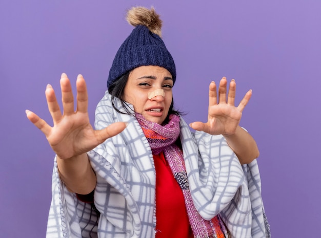 Unpleased young caucasian ill woman wearing winter hat and scarf with medical plaster on nose stretching out hands towards