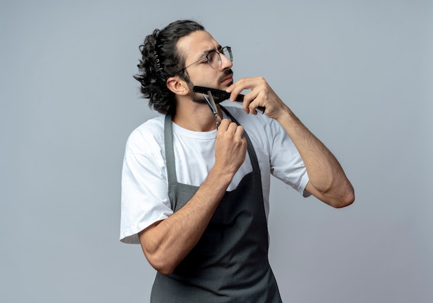 Unpleased young caucasian male barber wearing glasses and wavy hair band in uniform combing and cutting his beard with closed eyes isolated on white background with copy space