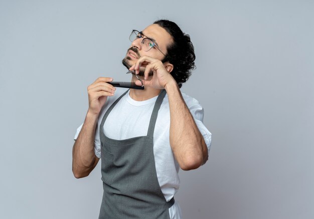 Unpleased young caucasian male barber wearing glasses and wavy hair band in uniform cutting and combing his beard looking straight isolated on white background with copy space