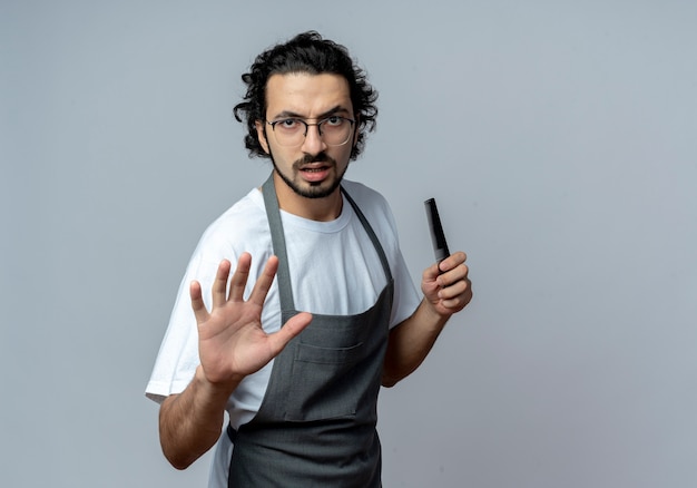 Barbiere maschio caucasico giovane dispiaciuto con gli occhiali e fascia per capelli ondulati in uniforme che tiene il pettine e allungando la mano alla fermata gesticolare della macchina fotografica isolata su fondo bianco con lo spazio della copia