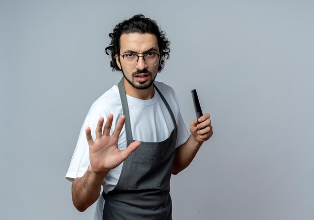 Unpleased young caucasian male barber wearing glasses and wavy hair band in uniform holding comb and stretching out hand at camera gesturing stop isolated on white background with copy space