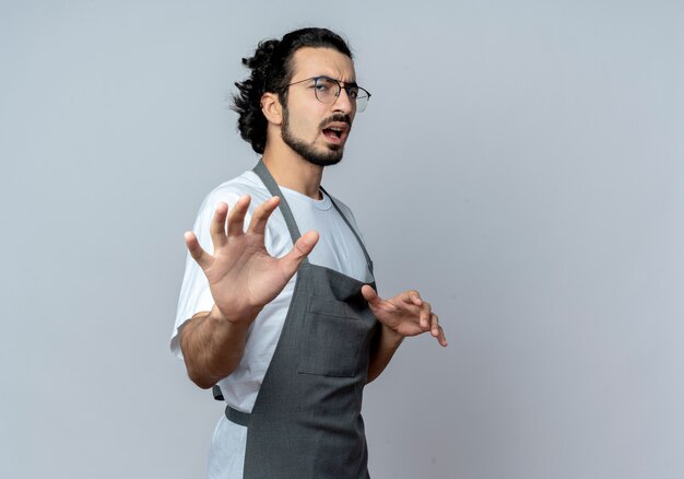 Unpleased young caucasian male barber wearing glasses and wavy hair band in uniform standing in profile view stretching out hand at camera gesturing no isolated on white background with copy space