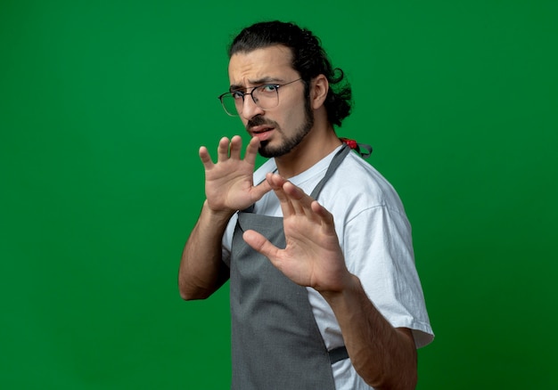 Unpleased young caucasian male barber wearing uniform and glasses standing in profile view stretching hand out at camera and keeping another one in air isolated on green background with copy space