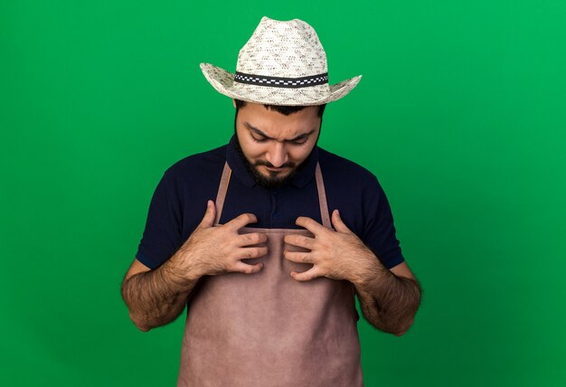 Scontento giovane maschio caucasico giardiniere che indossa cappello da giardinaggio mette le mani sul petto e guarda in basso isolato sulla parete verde con spazio di copia