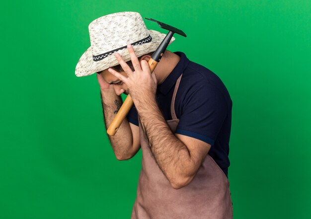 unpleased young caucasian male gardener wearing gardening hat puts hands on head holding rake isolated on green wall with copy space