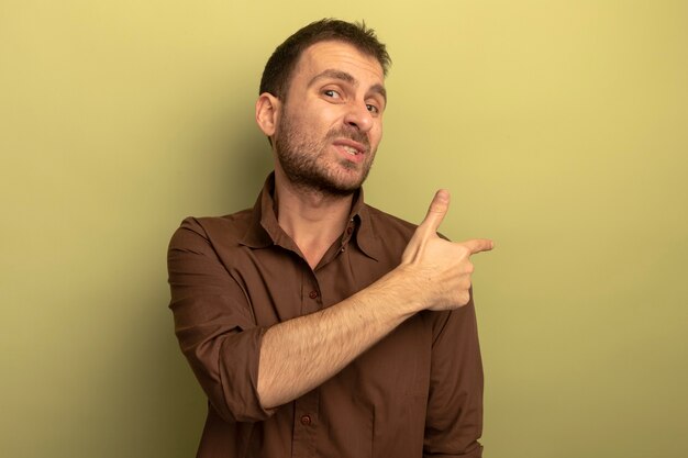 Недовольный молодой кавказский человек смотрит в камеру, указывая на сторону, изолированную на оливково-зеленом фоне с копией пространства