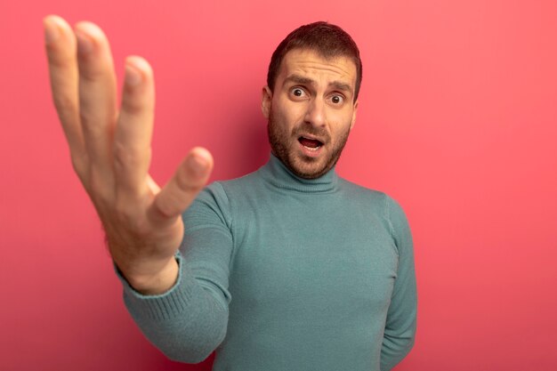 Unpleased young caucasian man looking at camera stretching out hand towards camera isolated on crimson background with copy space