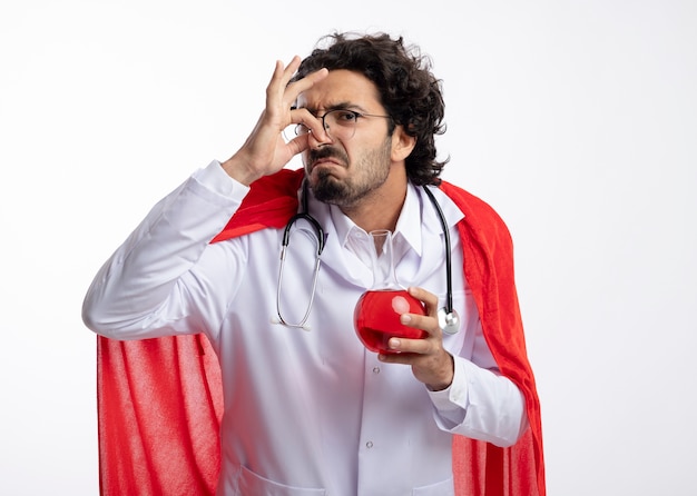 Unpleased young caucasian man in optical glasses wearing doctor uniform with red cloak and with stethoscope around neck closes nose and holds red chemical liquid in glass flask  on white wall