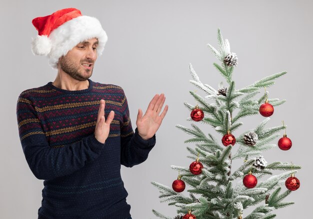 Giovane uomo caucasico dispiaciuto che indossa il cappello di natale in piedi vicino all'albero di natale guardandolo facendo gesto di rifiuto isolato su sfondo bianco