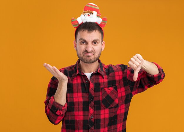 Unpleased young caucasian man wearing santa claus headband looking at camera showing empty hand and thumb down isolated on orange background