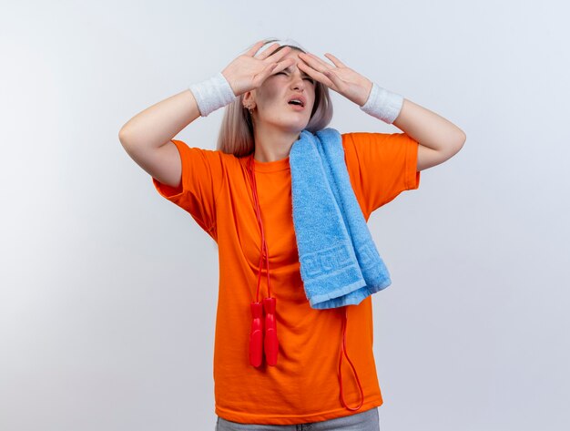 Unpleased young caucasian sporty girl with braces and with jumping rope around neck wearing headband and wristbands holding towel on shoulder puts hands on forehead  on white wall