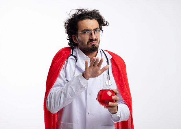 Недовольный молодой кавказский супергерой в оптических очках, одетый в форму доктора, красный плащ и со стетоскопом на шее, держит руку над красной химической жидкостью в стеклянной колбе