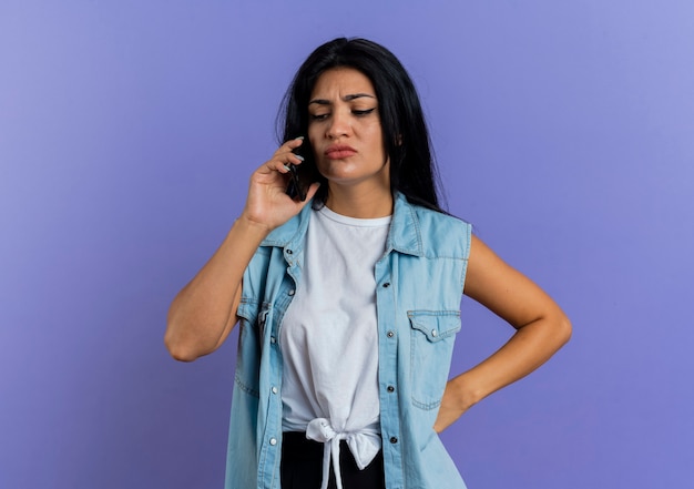 Unpleased young caucasian woman talks on phone and puts hand on waist isolated on purple background with copy space
