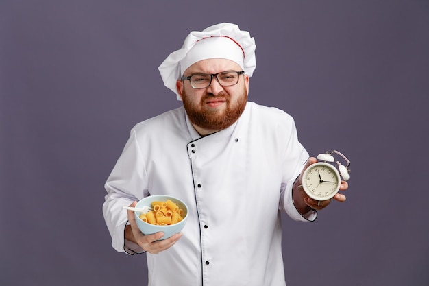 Бесплатное фото Недовольный молодой шеф-повар в очках, униформе и кепке, держащий миску макарон с ложкой в ней, смотрит в камеру, показывающую будильник, изолированный на фиолетовом фоне
