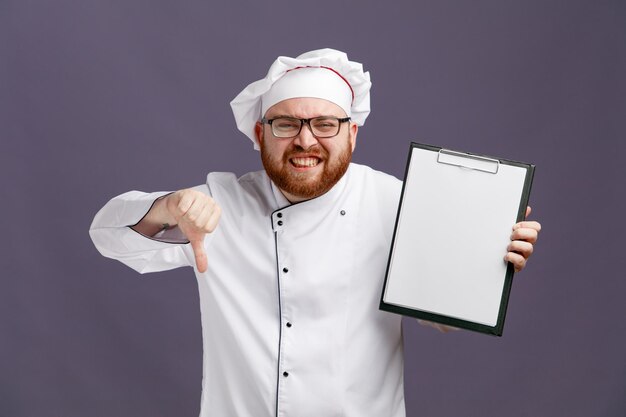 Unpleased young chef wearing glasses uniform and cap showing clipboard looking at camera showing thumb down isolated on purple background