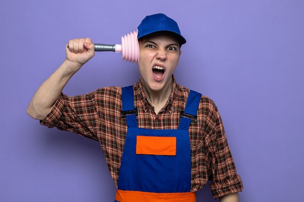 Foto gratuita giovane ragazzo delle pulizie scontento che indossa l'uniforme e il cappuccio che tiene lo stantuffo sulla testa