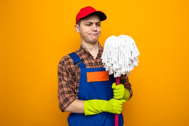 Unpleased young cleaning guy wearing uniform and cap with gloves holding and looking at mop 
