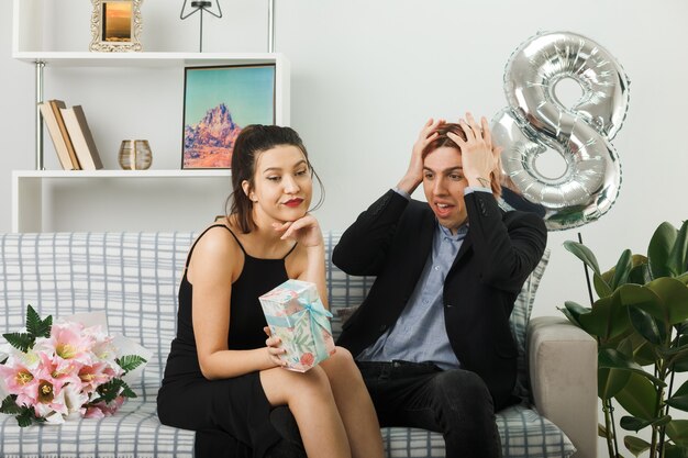 Unpleased young couple on happy women day holding present guy putting hands on head sitting on sofa in living room