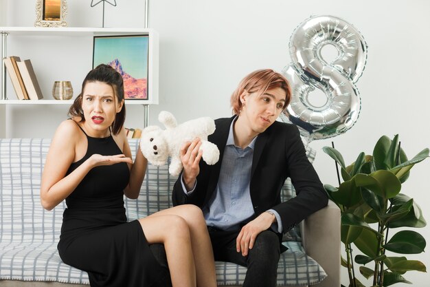 Недовольная молодая пара в счастливый женский день с плюшевым мишкой, сидящим на диване в гостиной
