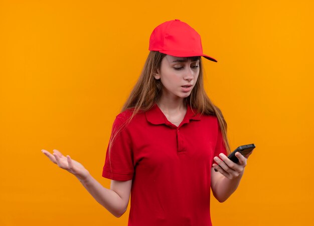 Unpleased young delivery girl in red uniform holding mobile phone looking at it showing empty hand on isolated orange wall