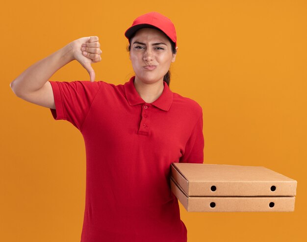 Unpleased young delivery girl wearing uniform and cap holding pizza boxes showing thumb down isolated on orange wall
