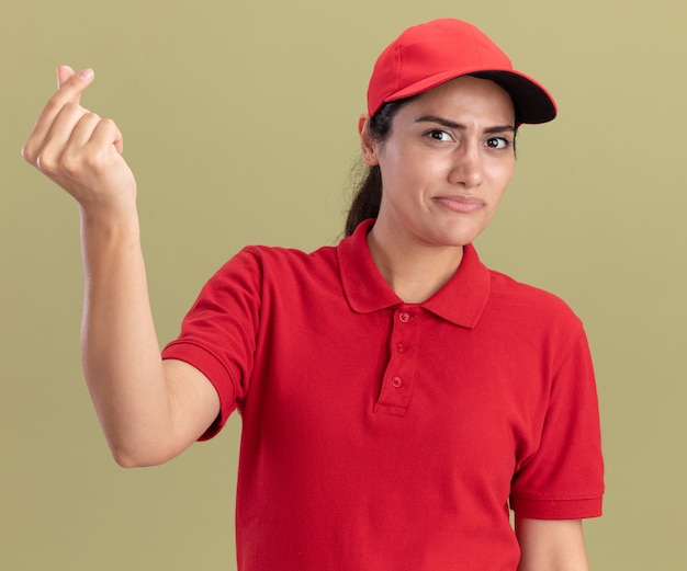 Недовольная молодая доставщица в униформе с кепкой, показывающая жест кончика, изолирована на оливково-зеленой стене