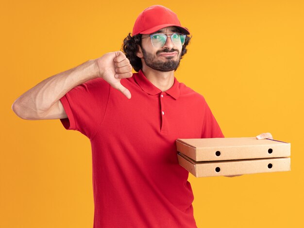 Unpleased young delivery man in red uniform and cap wearing glasses holding pizza packages looking at front showing thumb down isolated on orange wall