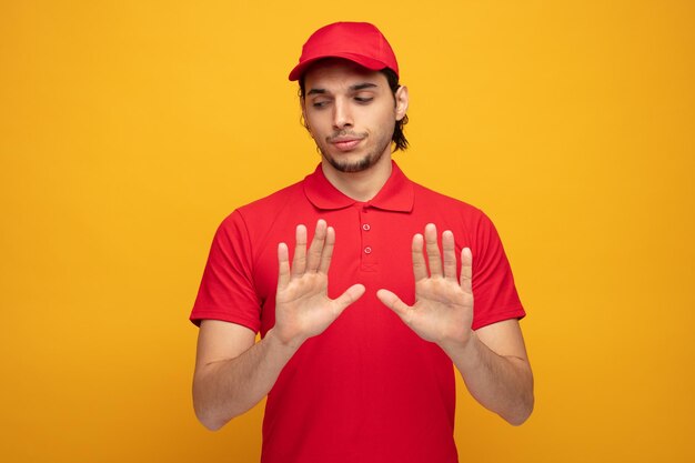 unpleased young delivery man wearing uniform and cap looking at side showing stop gesture with both hands isolated on yellow background