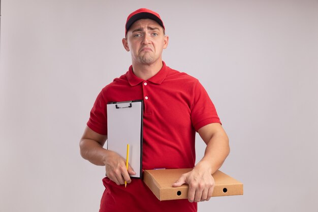 Unpleased young delivery man wearing uniform with cap holding clipboard with pizza box isolated on white wall