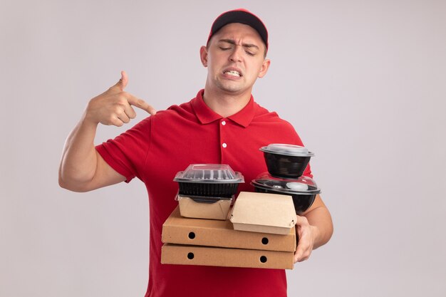 Unpleased young delivery man wearing uniform with cap holding and points at food containers on pizza boxes isolated on white wall