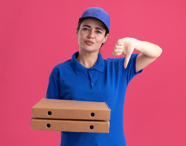 Giovane donna delle consegne scontenta in uniforme e berretto che tiene i pacchetti di pizza che mostra il pollice verso il basso isolato sul muro rosa