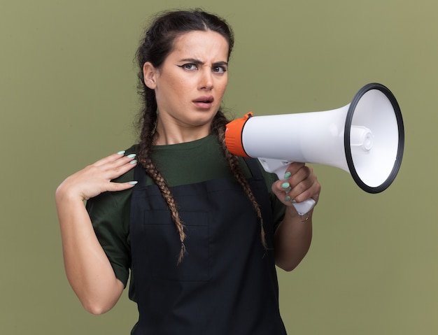 Foto gratuita il giovane barbiere femminile scontento in uniforme parla sull'altoparlante mettendo la mano sulla spalla isolata sulla parete verde oliva