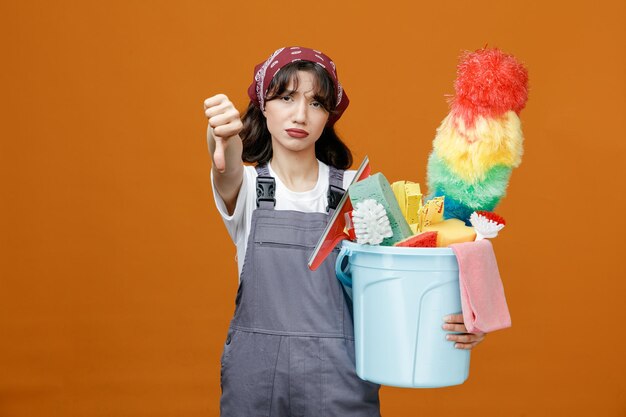 Недовольная молодая женщина-уборщица в униформе и бандане, держащая ведро чистящих инструментов, смотрит в камеру, протягивая руку, показывая большой палец вниз на оранжевом фоне