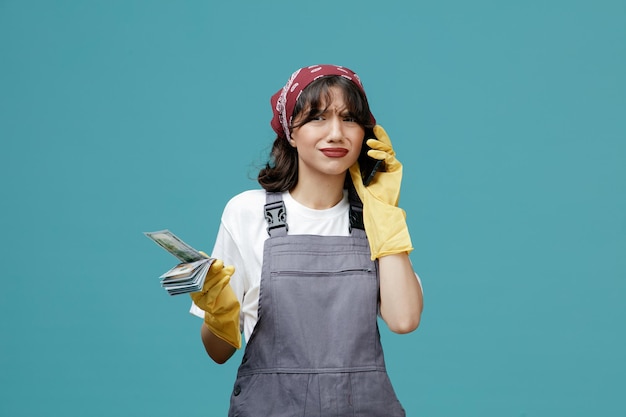 Free photo unpleased young female cleaner wearing uniform bandana and rubber gloves holding money looking at camera while talking on phone isolated on blue background