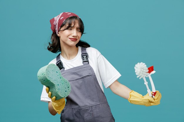 制服のバンダナとゴム手袋を身に着けている不機嫌な若い女性クリーナーは、青い背景に分離されたブラシを見て、スポンジストレッチトイレブラシを保持しています