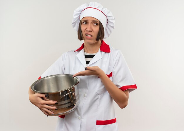 Unpleased young female cook in chef uniform holding and pointing at pot looking at side isolated on white background with copy space