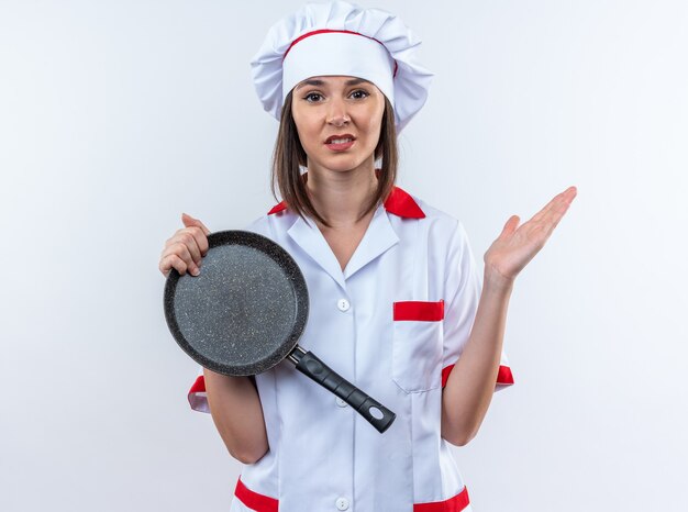 白い壁に分離された手を広げてフライパンを保持しているシェフの制服を着て不機嫌な若い女性料理人