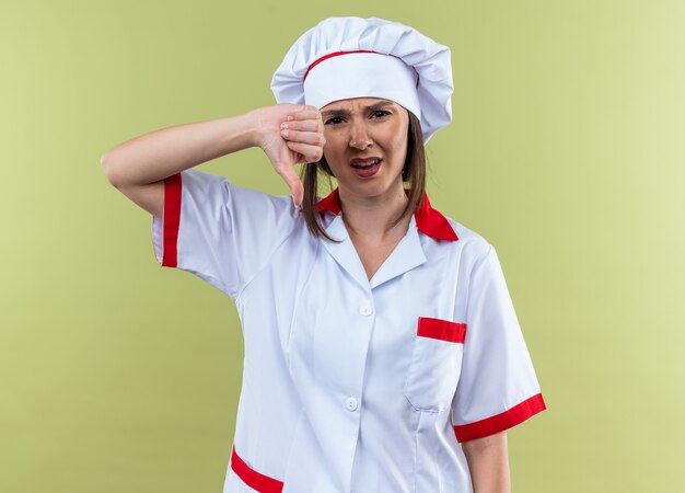 unpleased young female cook wearing chef uniform showing thumb down isolated on olive green wall