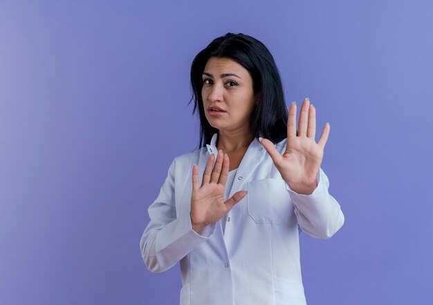コピースペースのある紫色の壁に隔離されたジェスチャーをしない医療ローブを身に着けている不機嫌な若い女性医師