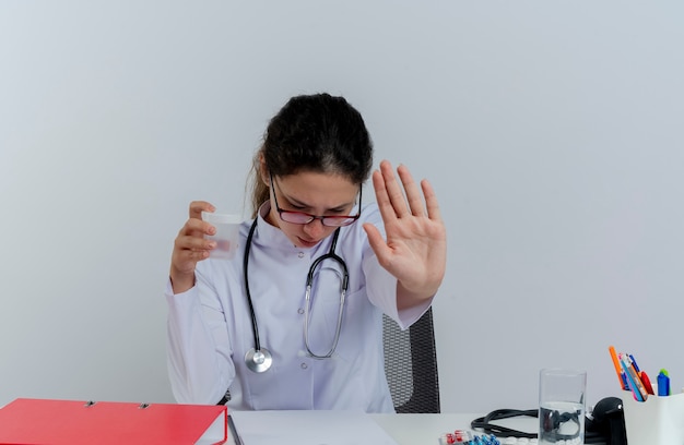 Unpleased young female doctor wearing medical robe and stethoscope and glasses sitting at desk with medical tools holding medical beaker looking down doing stop gesture isolated