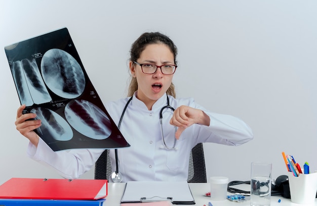 Недовольная молодая женщина-врач в медицинском халате, стетоскопе и очках, сидящая за столом с медицинскими инструментами, держащая рентгеновский снимок, показывающая изолированный палец вниз